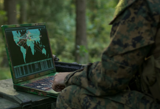 Military Operation in Action, Soldiers Using Military Grade Laptop Use Military Industrial Complex Hardware for Accomplishing International Mission. In the Background Camouflaged Tent on the Forest.